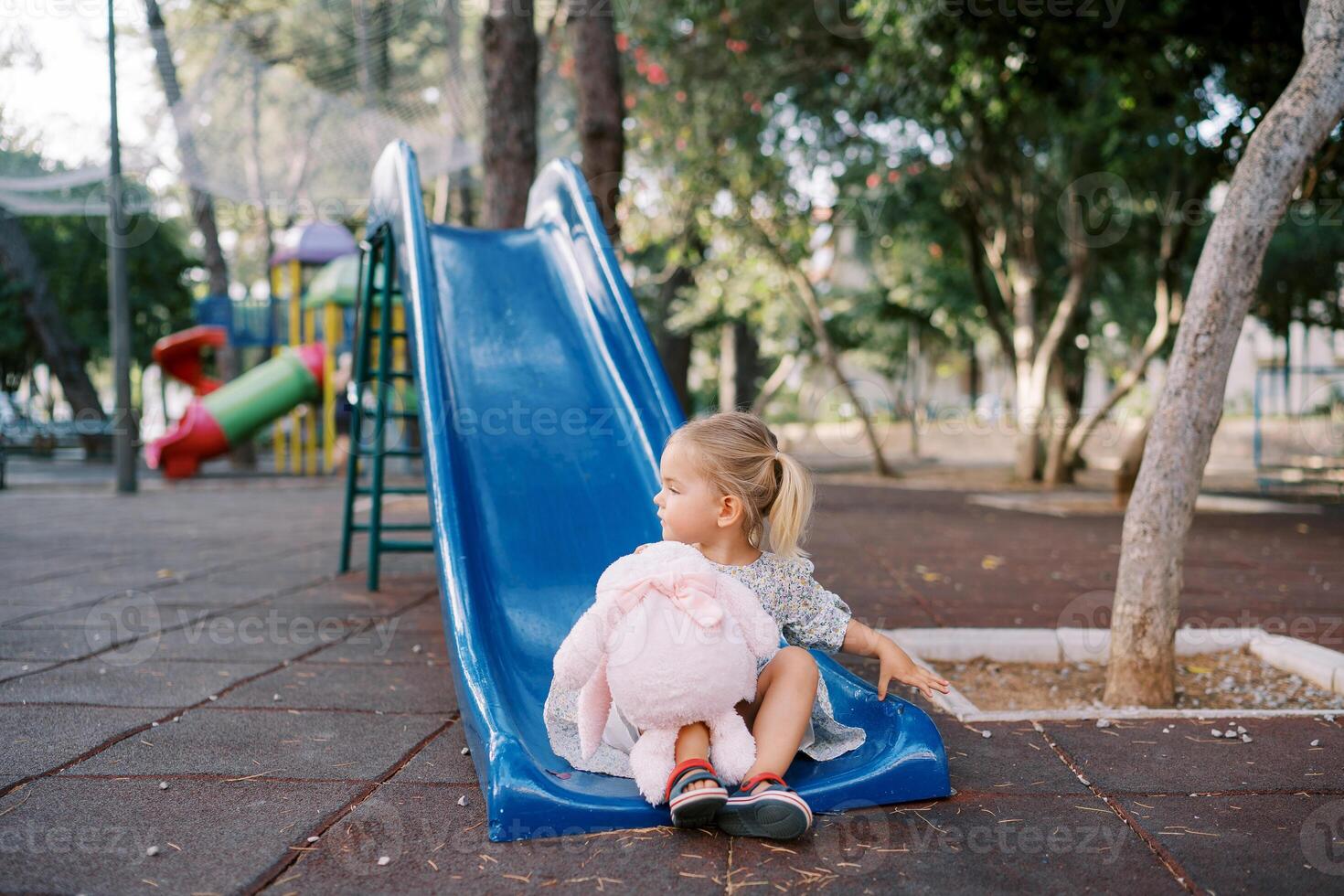 Little girl with a pink plush bunny sits on a slide and looks away photo
