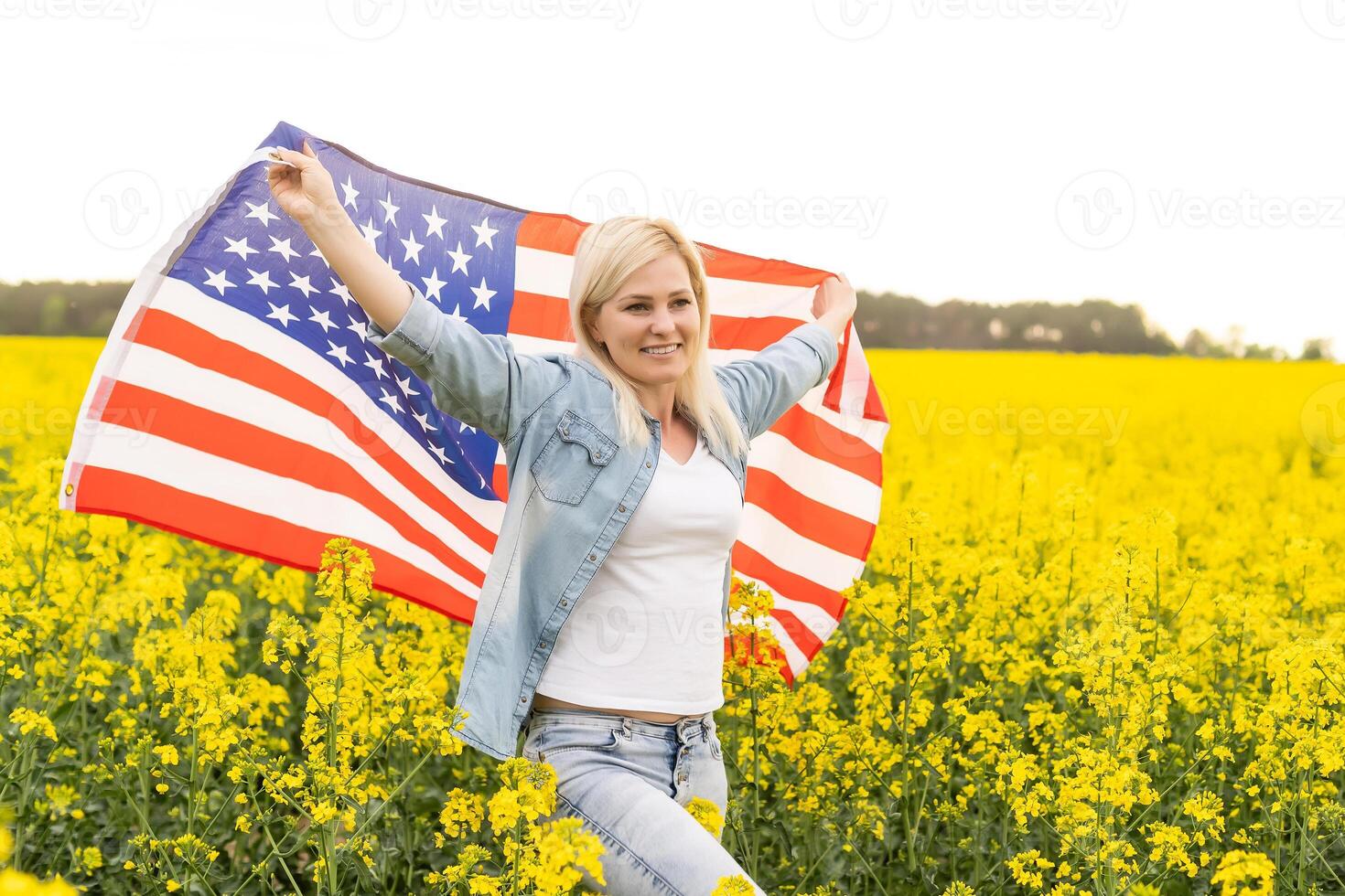 attractive woman holding an American flag in the wind in a field of rapeseed. Summer landscape against the blue sky. Horizontal orientation. photo