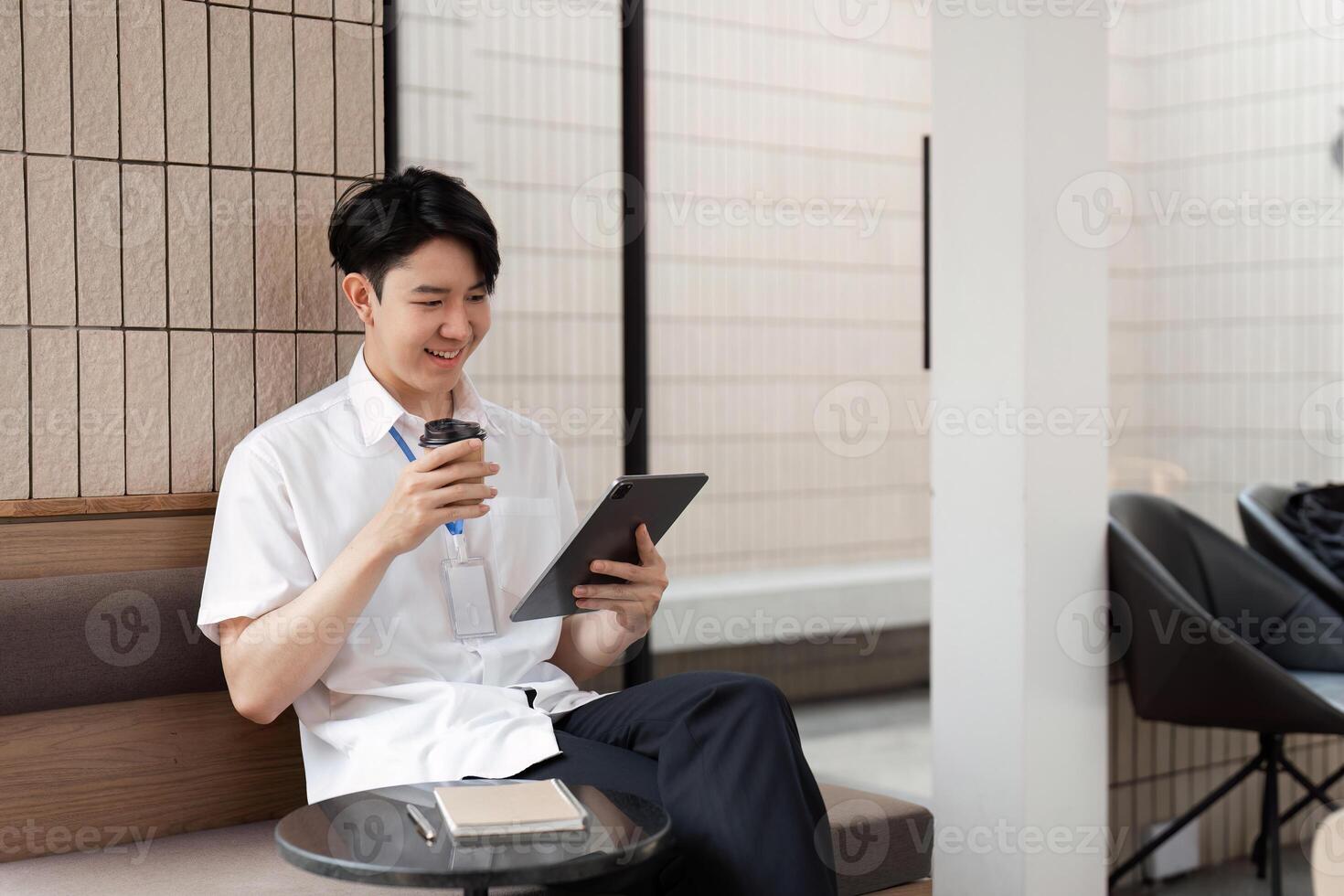 Young Asian male office worker smiling while using tablet in modern workspace, enjoying coffee and digital content. photo