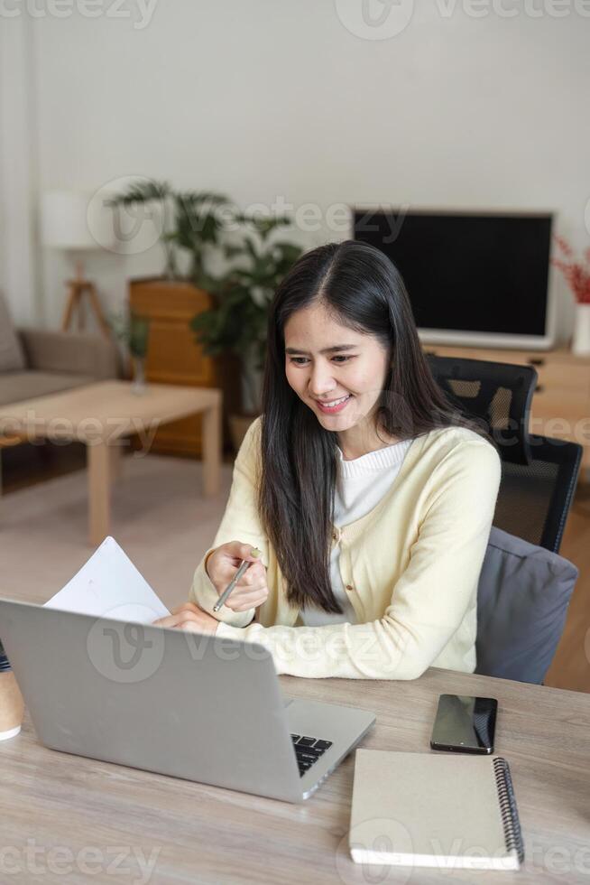 Smiling Asian woman working on a laptop while reviewing documents, showcasing a positive and productive home office atmosphere. photo