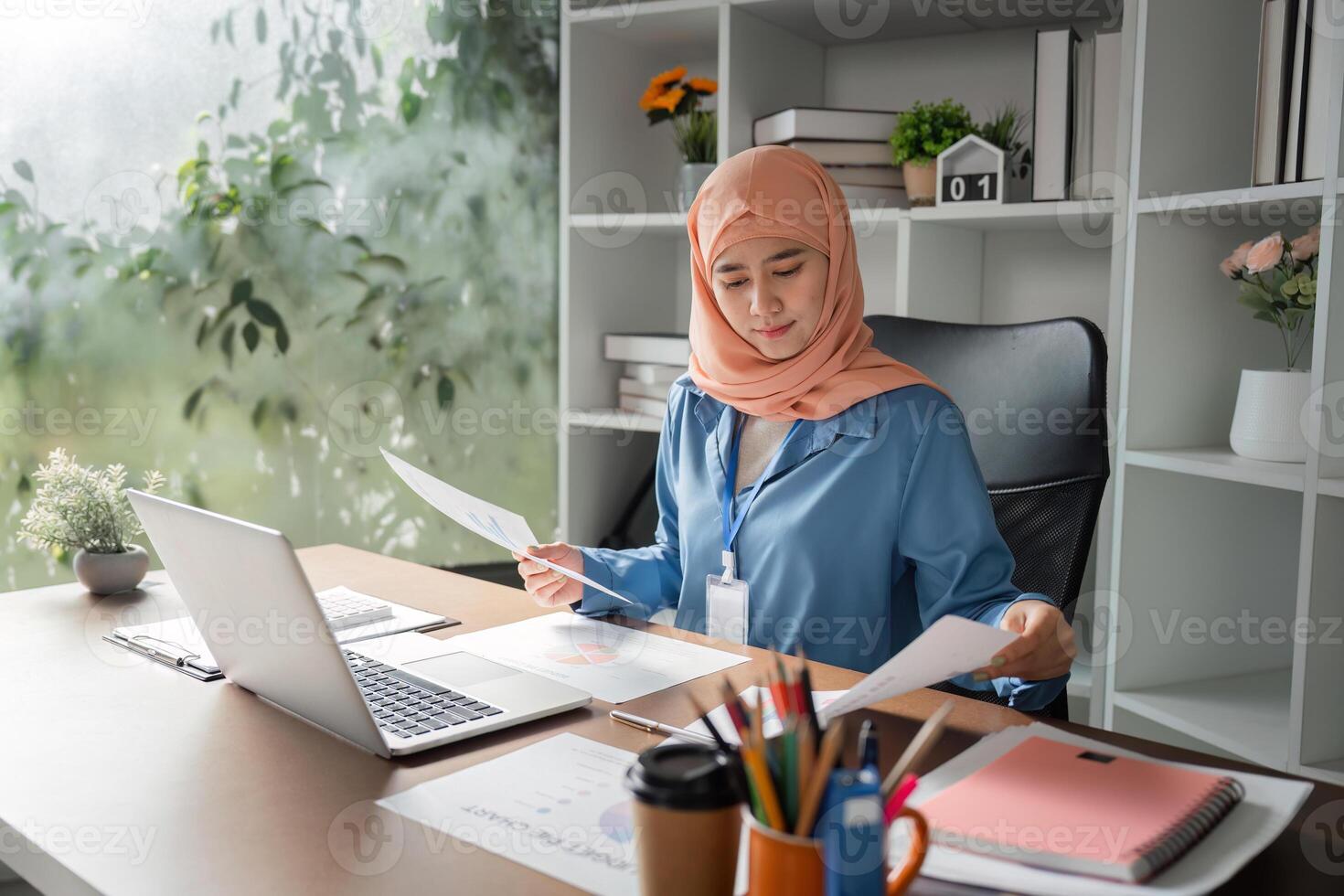 Muslim woman analyzing financial reports while sitting at a desk with a laptop. photo
