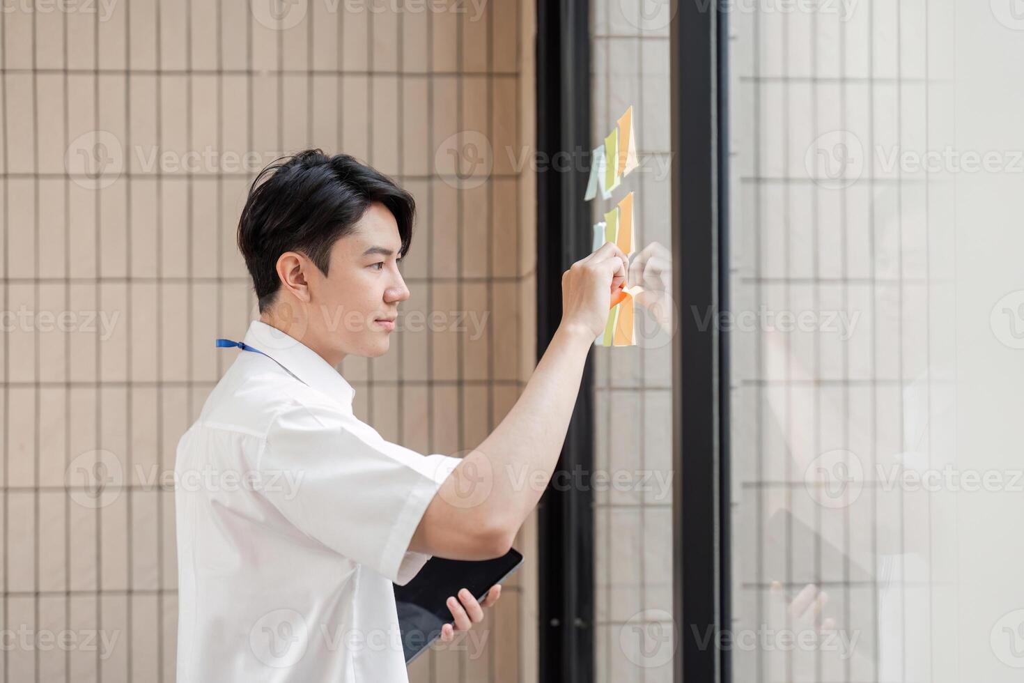 Young Asian male office worker organizing sticky notes on glass wall in modern workspace, focusing on planning and strategy. photo