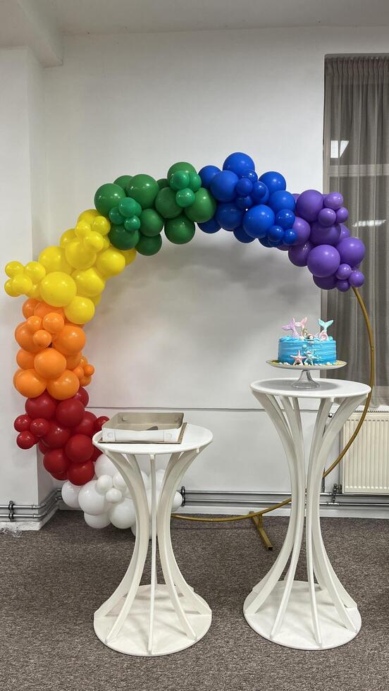 Mermaid themed birthday cake with mermaid tails, seashells, pearls, and a 6 candle. Party decorations include a balloon arch with blue, purple, yellow, and green balloons photo