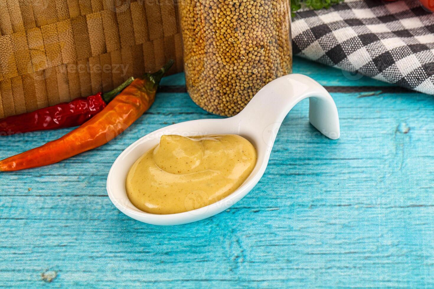 A spoon with mustard and a bowl of mustard on a wooden table photo