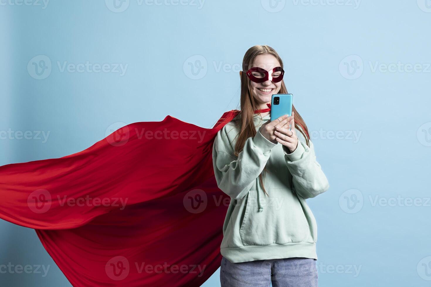 Portrait of upbeat teenager with mask sending messages on cellphone, portraying superhero, studio backdrop. Radiant hero character in costume texting friends on mobile phone photo