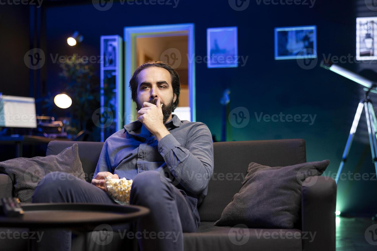 Lazy man enjoying leisure time at home watching TV, munching on popcorn. Couch potato in neon illuminated apartment, stuffing mouth with salty snack, entertained by television photo