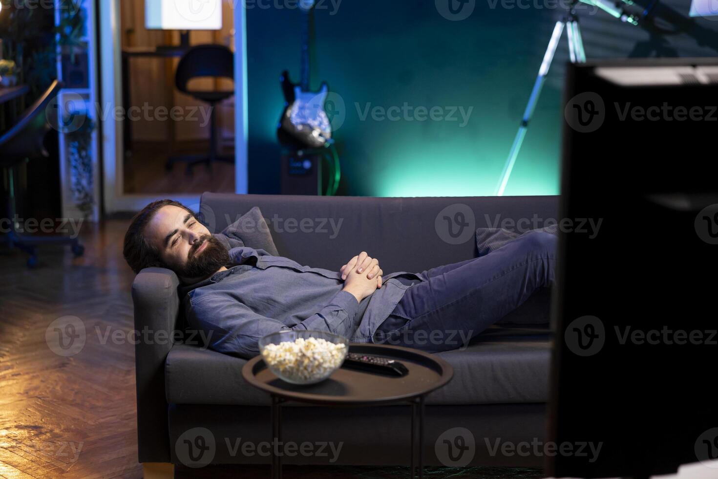 Smiling man watching feel good movies at home on TV, engrossed by riveting action, paying close attention to screen. Cheerful person relaxing in apartment lounged on couch, enjoying film and snacks photo