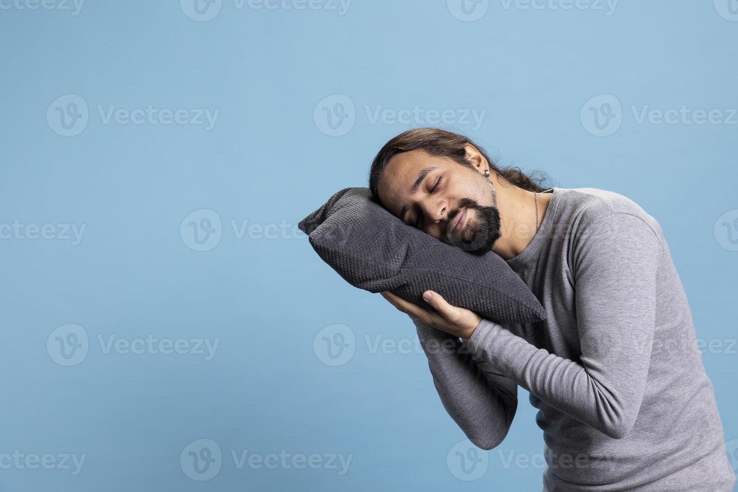 Tired fatigued person sleeping on a grey pillow against blue background, acting exhausted and taking a nap in front of the camera. Young adult being sleepy and falling asleep standing. photo