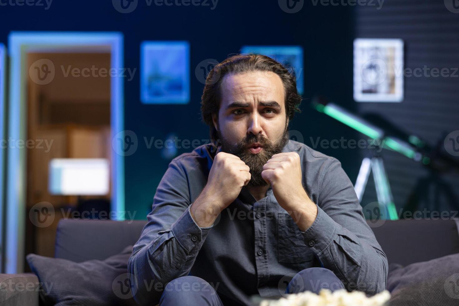 Man watching shows on TV, clenching punches, rooting for characters, paying close attention to screen. Man sitting on couch in apartment doing clasping hands gesture, hoping for happy ending photo