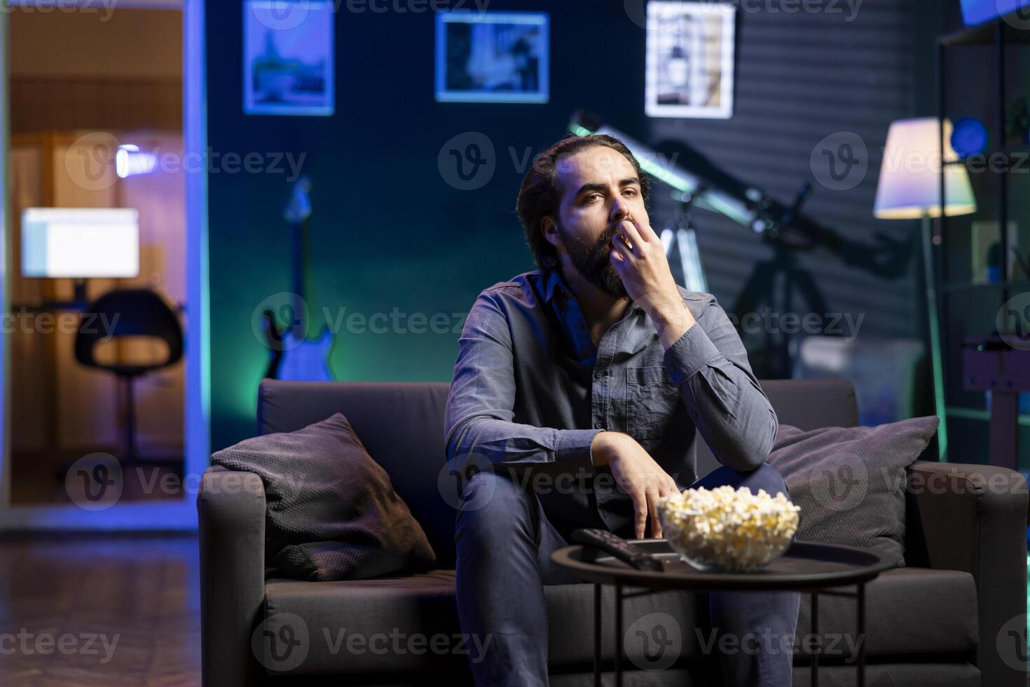 Man relaxing at home on couch, stuffing mouth with popcorn while watching movies on TV in apartment. Caucasian person eating snacks and streaming shows in neon lit living room photo