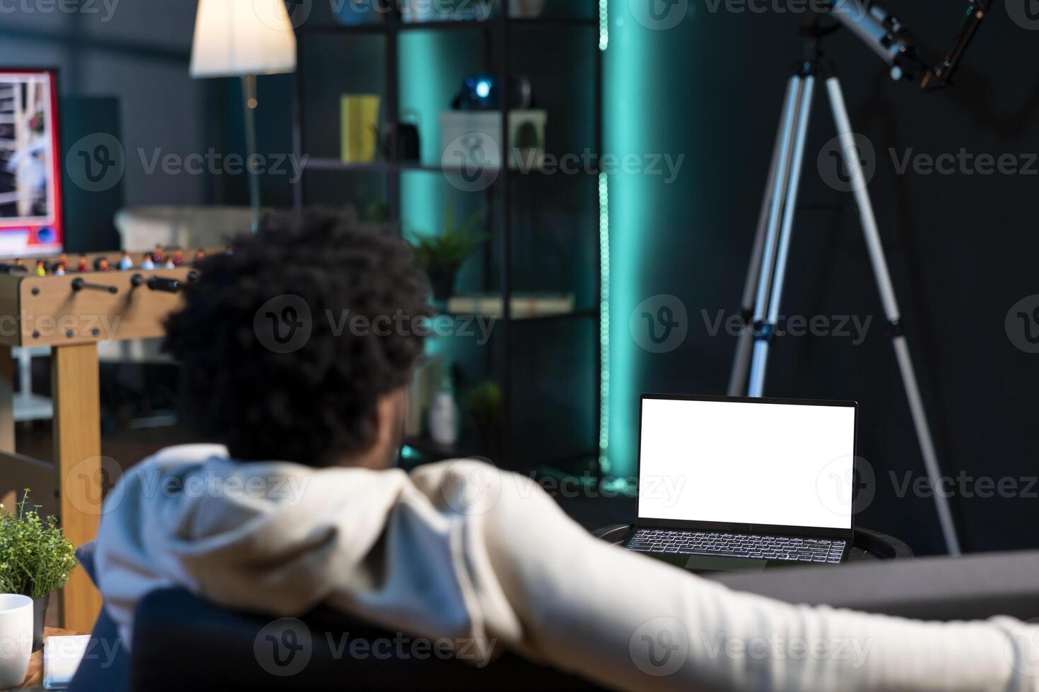 Man relaxing in living room by watching TV series on mockup laptop. BIPOC person enjoying leisure time in neon illuminated home, looking at multimedia content on isolated screen notebook photo