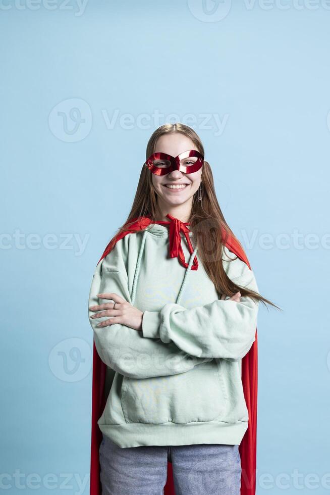 Portrait of happy teenager with mask portraying confident superhero wearing red cape, studio backdrop. Jolly heroic person posing as powerful hero character in costume, concealing identity photo