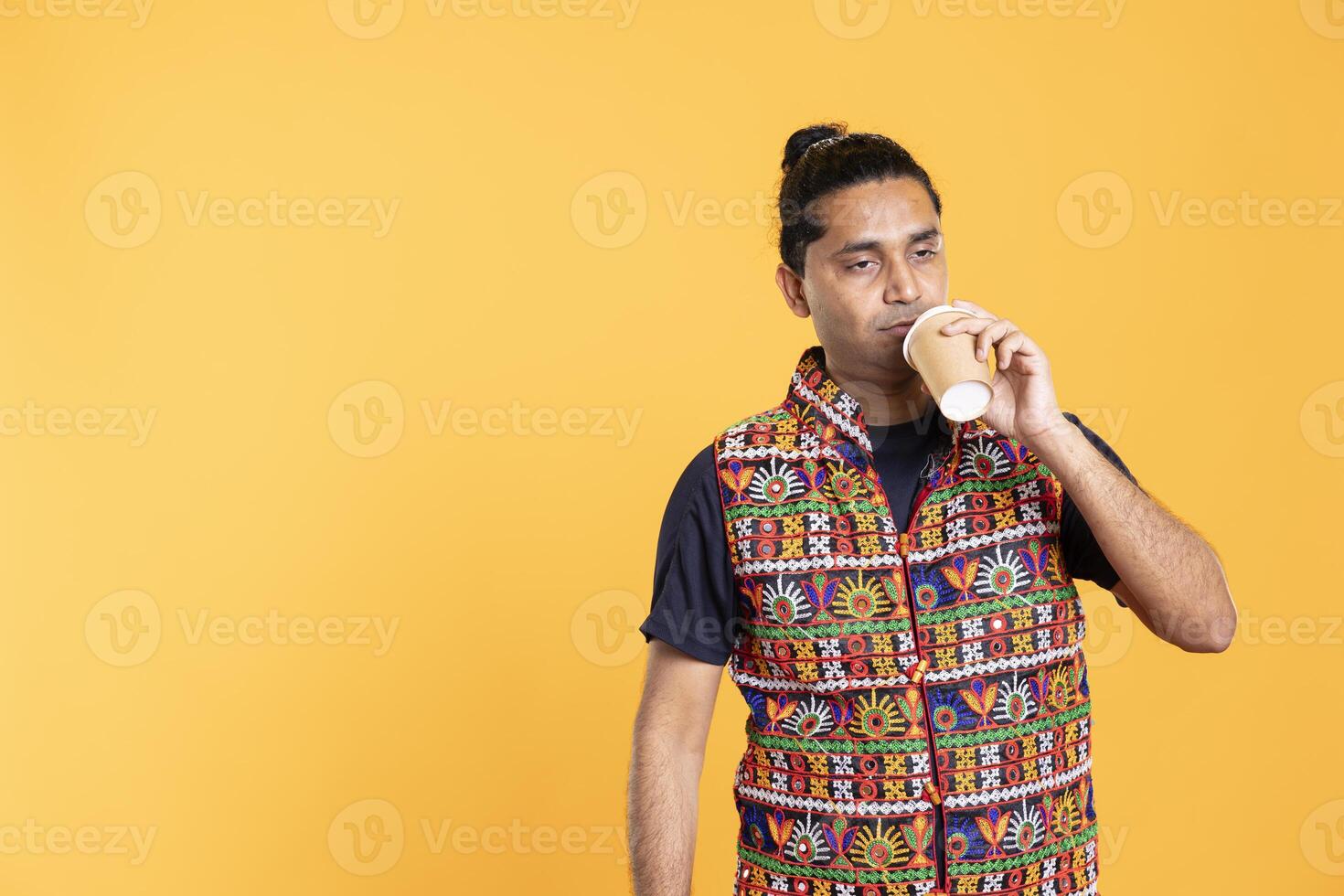 Tired indian man sipping fresh coffee from disposable paper cup early in the morning to wake up and be energized. Person drinking hot beverage from recycled takeaway cup, studio background photo