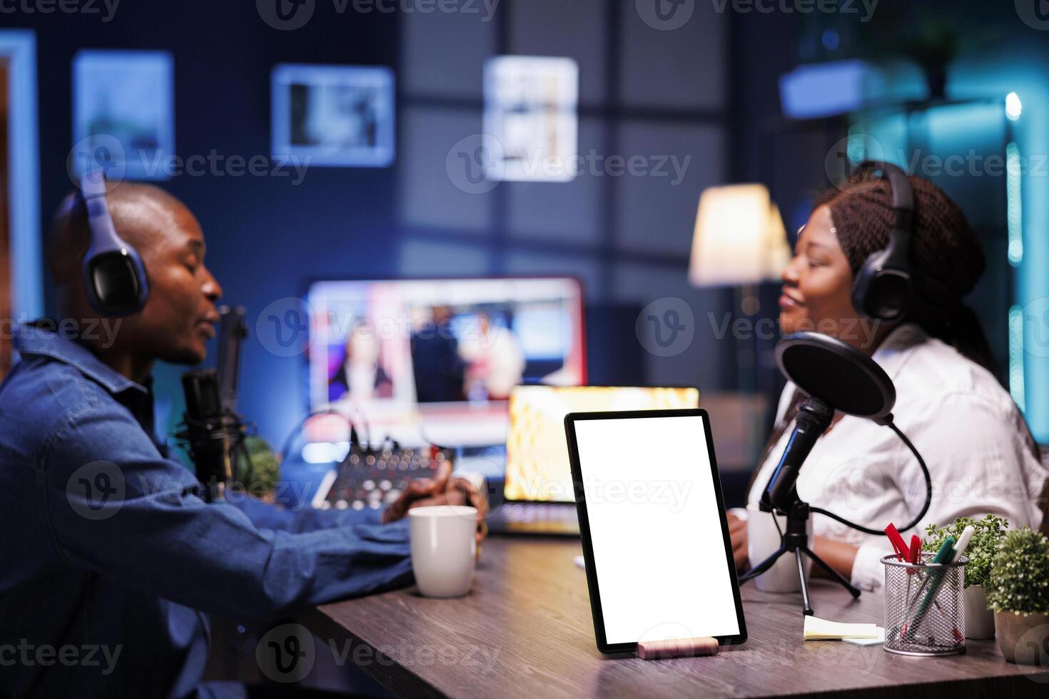 On desk, white screen with mockup template is displayed on tablet while African American vloggers are recording live podcast while wearing headsets. Digital device on table shows isolated chromakey. photo