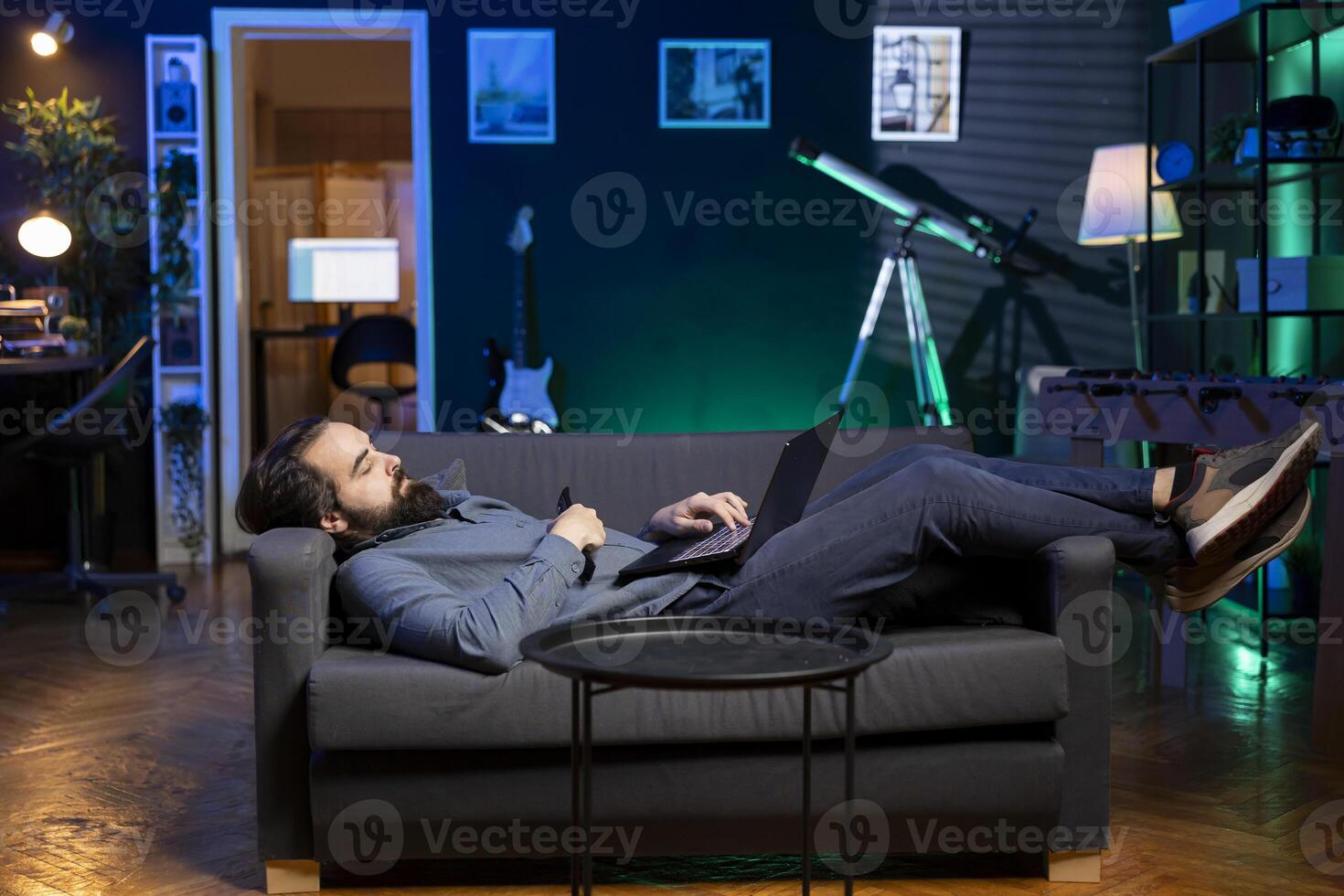 Homeowner bored at home laying down, using TV to watch movies on streaming services and typing on laptop. Man lounged on couch, mindlessly consuming entertainment with notebook in lap photo