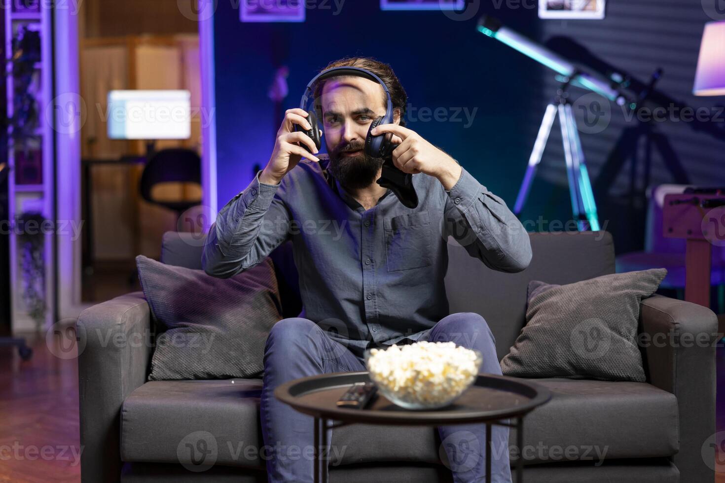 Smiling man putting headset on to play videogames in neon lit apartment, holding controller. Gamer participating in game using console system and headphones for immersive experience photo