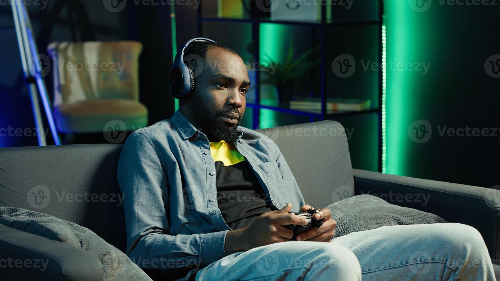 Tired gamer in RGB lit home late at night gaming with gamepad. Man sitting on sofa in neon illuminated apartment, playing videogames on console using controller, looking exhausted, camera A photo