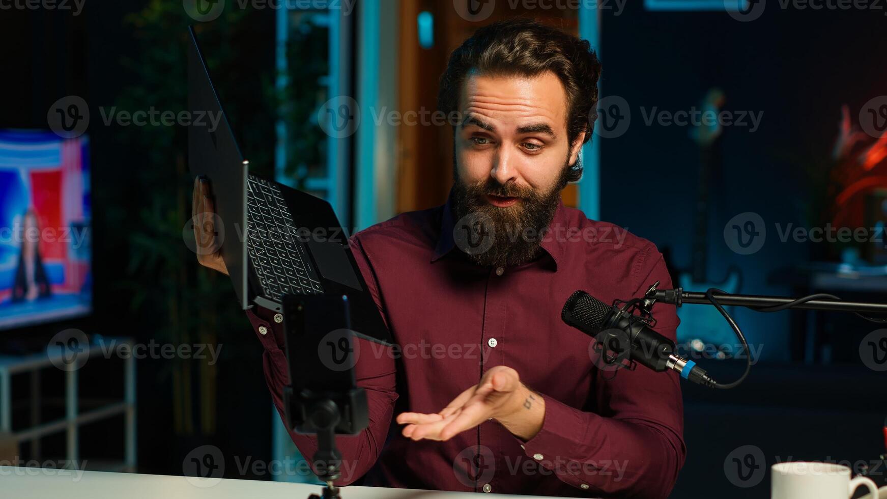Man in studio endorsing laptop from brand sponsoring , filming with smartphone on tripod. Tech expert does influencer marketing by promoting notebook, recording with phone, camera A photo