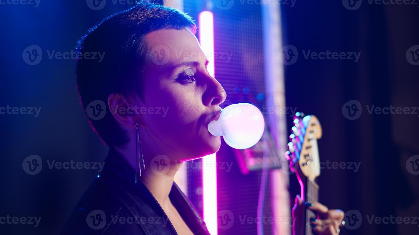 Cool grunge artist blowing bubbles with bubble gum flavor in studio, chewing gum and acting tough. Female musician rocker with leather jacket performs alternative music. Camera A. photo