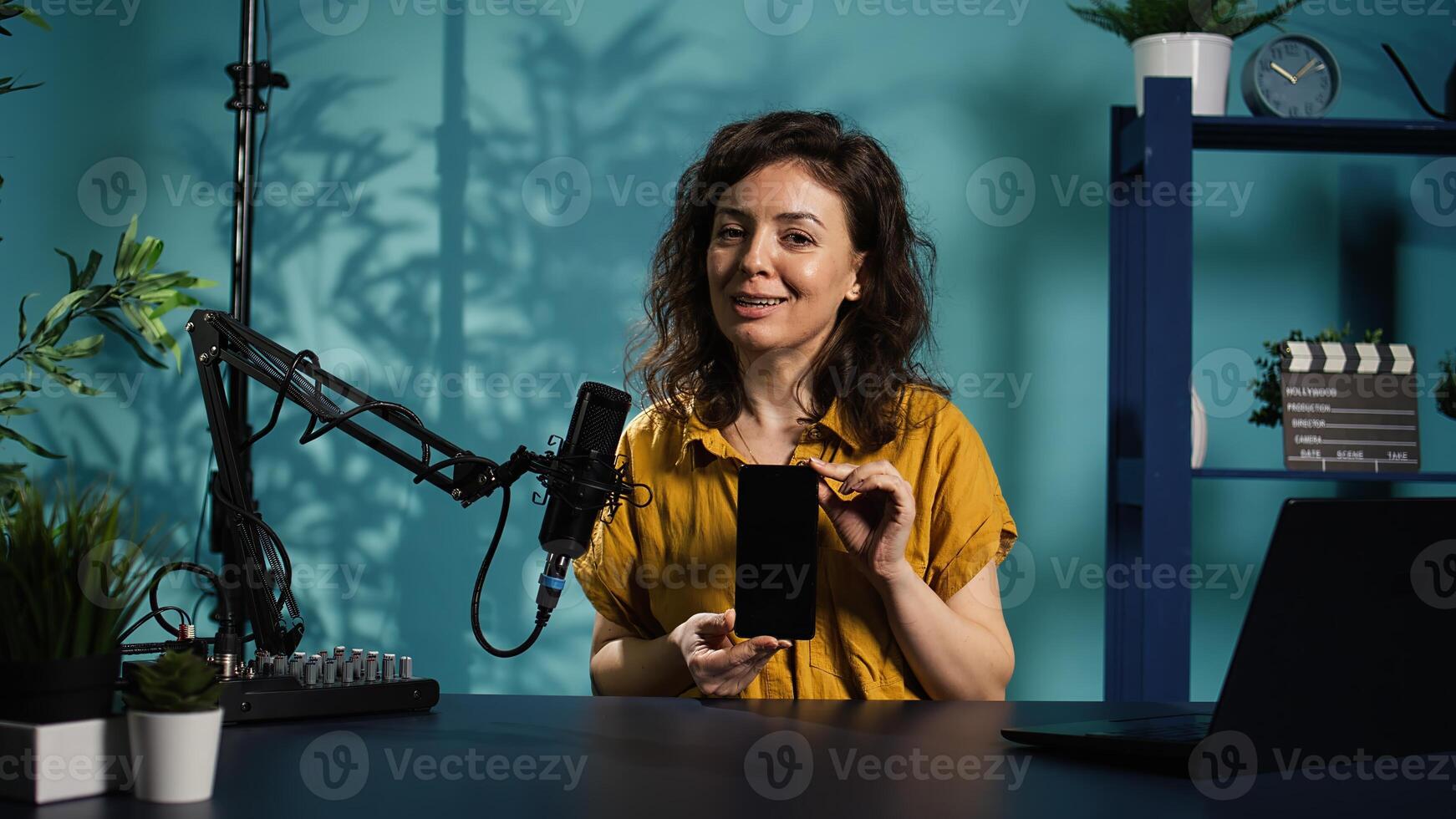 Cheerful entertainer presenting mobile phone from brand sponsoring , urging viewers to purchase it. Smiling woman does influencer marketing by advertising smartphone to audience, camera B photo