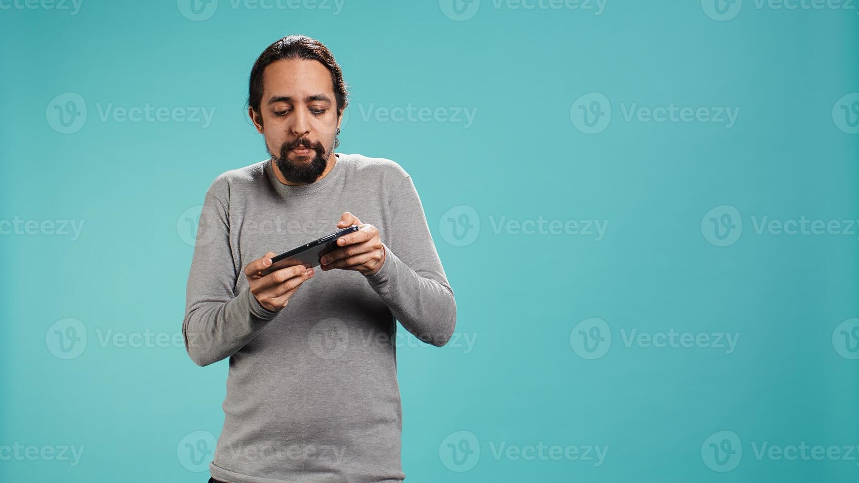 Focused man plays intense videogame on mobile phone, spamming attack buttons on touchscreen. Gamer participates in gaming competition, isolated over studio background, camera B photo