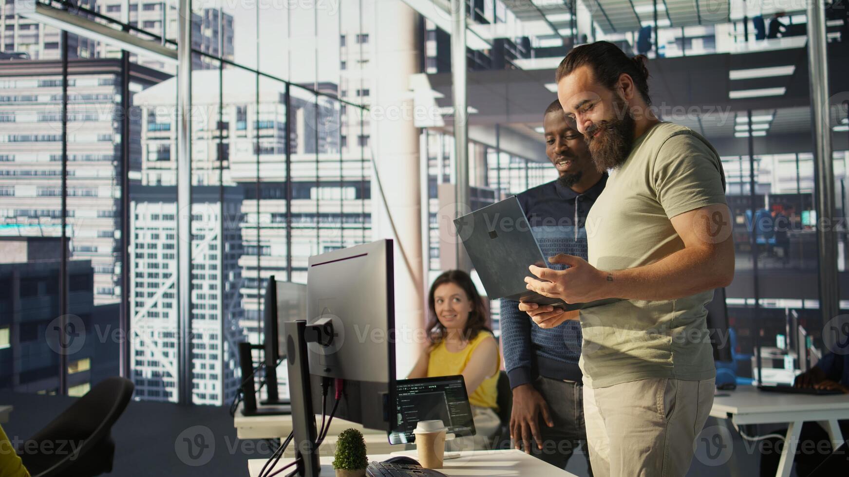 Cheerful admin holding laptop chatting with colleagues while working with machine learning tech. Man transforming data into predictive models using notebook and talking with coworkers, camera B photo