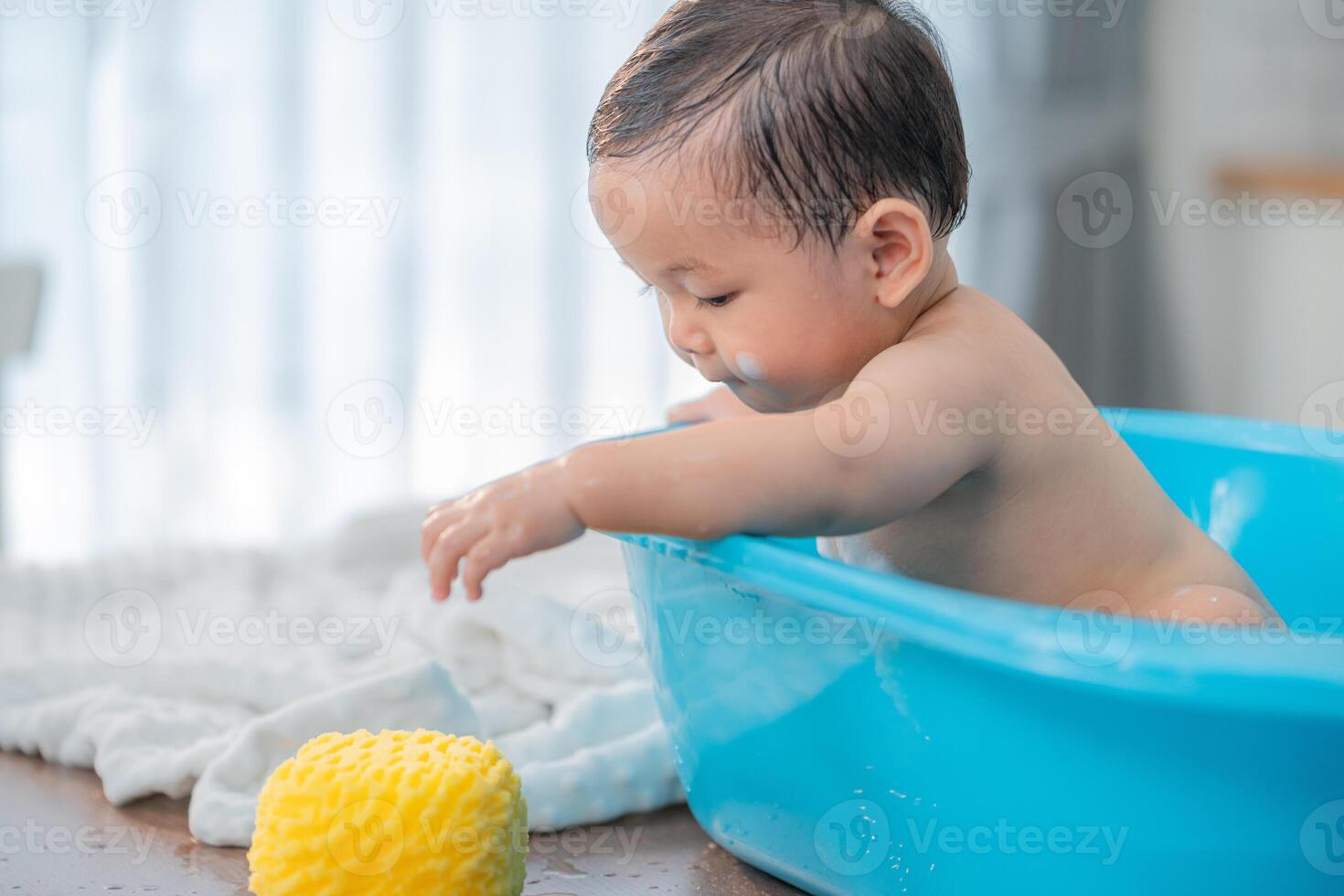 Baby enjoying bath time in a blue tub with a yellow sponge in a bright indoor setting during the day photo