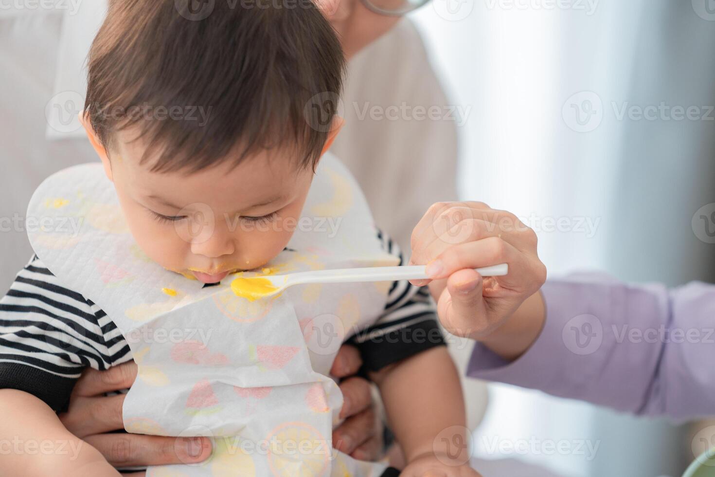 Baby enjoying mealtime as caregiver feeds with a spoon in a bright indoor setting photo