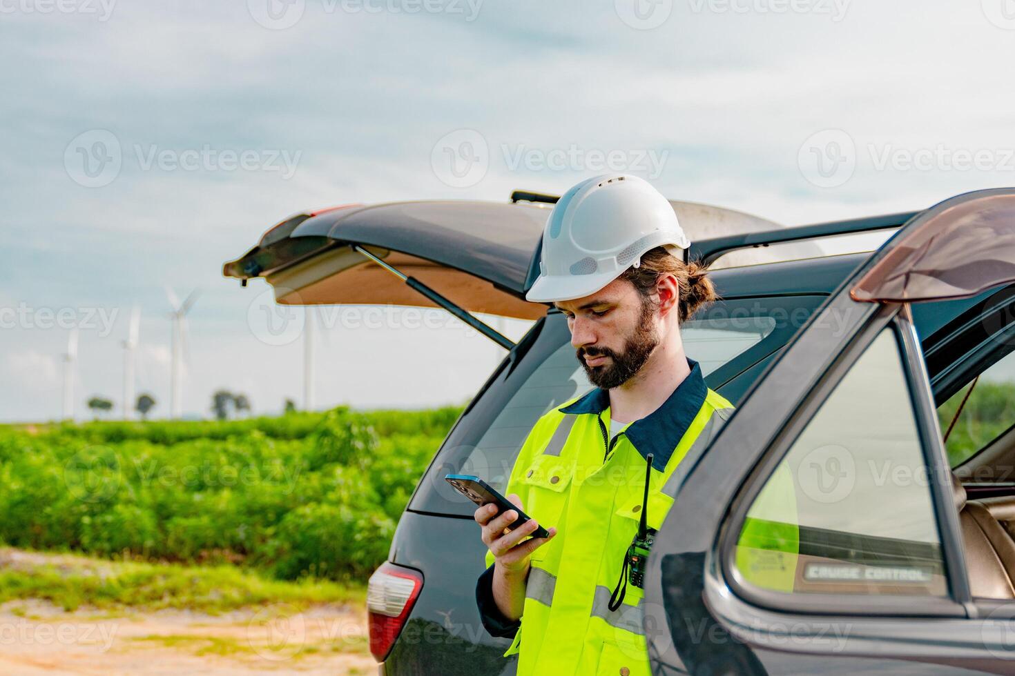 Worker in safety gear checks smartphone in rural area surrounded by wind turbines and green landscape during daylight hours photo