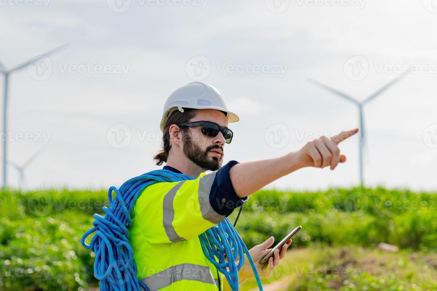 Worker directs team in renewable energy field while wearing safety gear and holding a smartphone photo