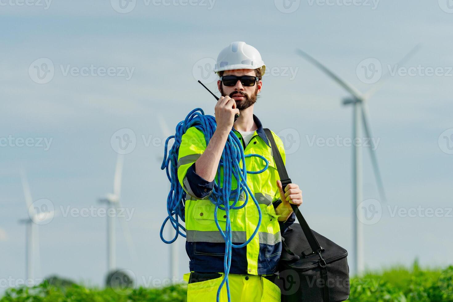 Worker in safety gear communicates while standing near wind turbines in a green field during the day photo