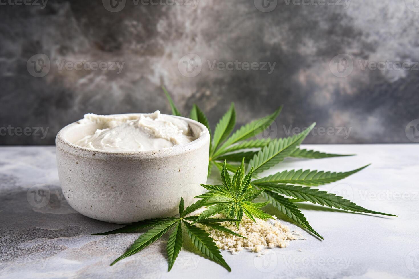 close-up of a white jar of organic cream surrounded by green cannabis leaves standing on table on a grey concrete background photo