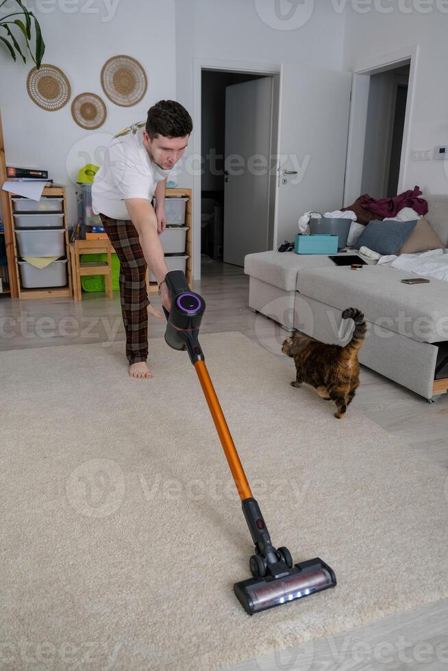 Man cleaning carpet with a vacuum while a cat watches, daily home routine photo