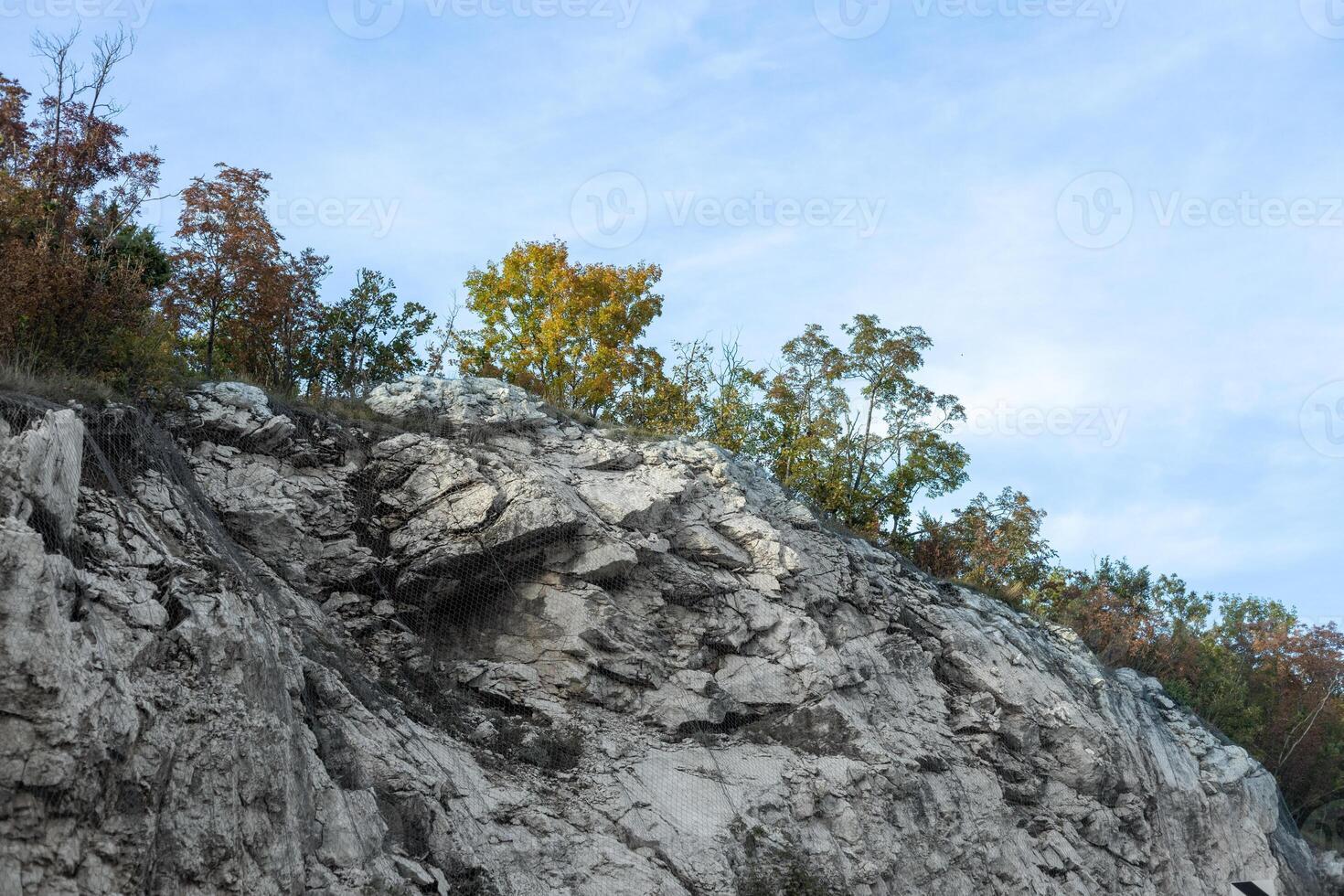 Rocky cliff with patches of green and autumn trees under a partly cloudy blue sky depicting natural erosion and geology. photo