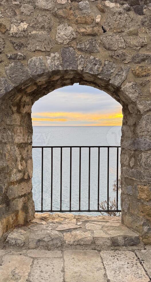 A rustic stone archway frames a view of calm blue waters and a golden sunset, with a black iron railing in the foreground. High quality photo