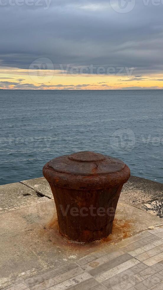 A large rusted mooring bollard on a stone pier with calm sea waters and a dramatic sunset sky in the background. photo