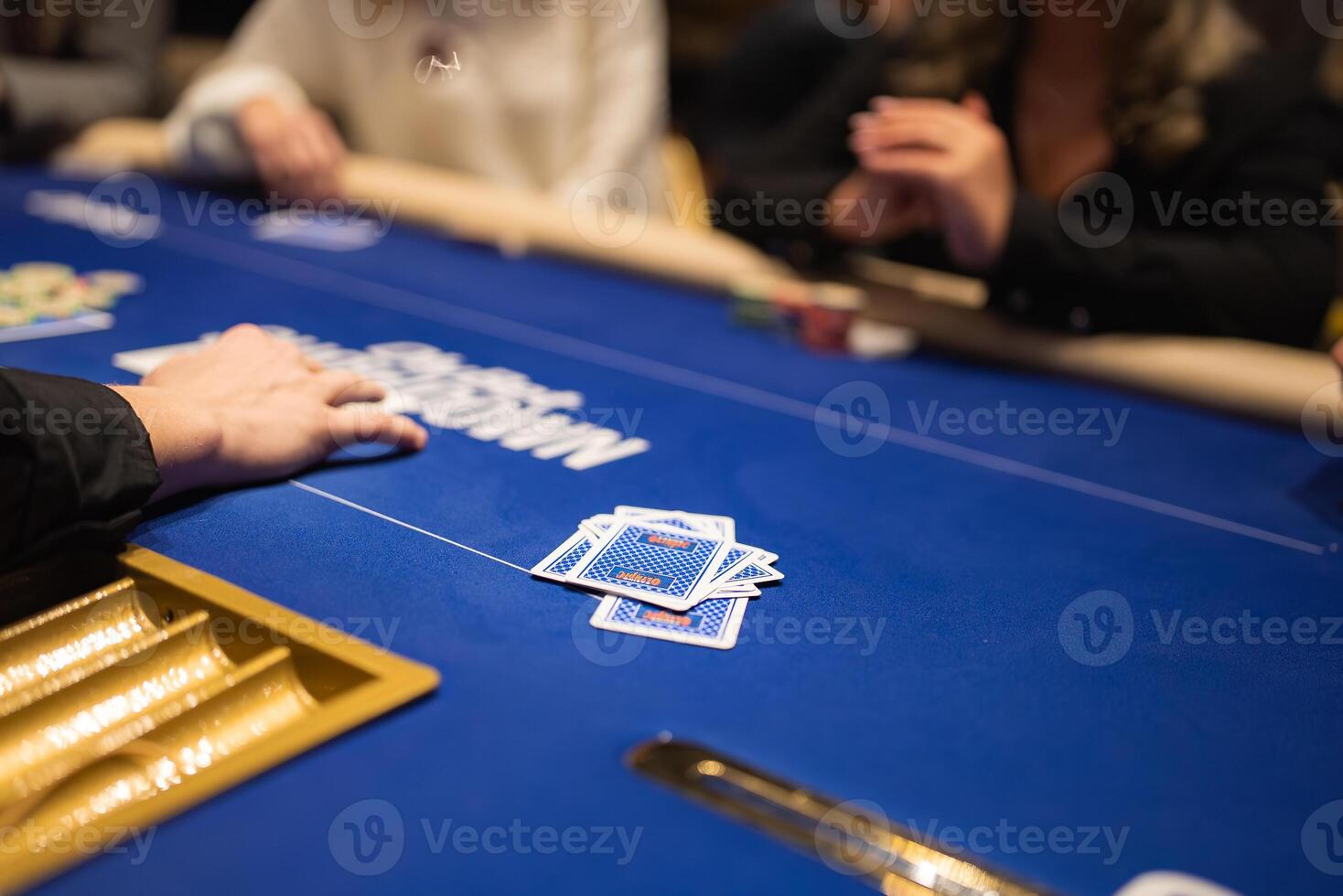 A poker game in progress on a blue felt table with visible branding, a gold card slot, chips, and hands reaching toward face down cards. photo