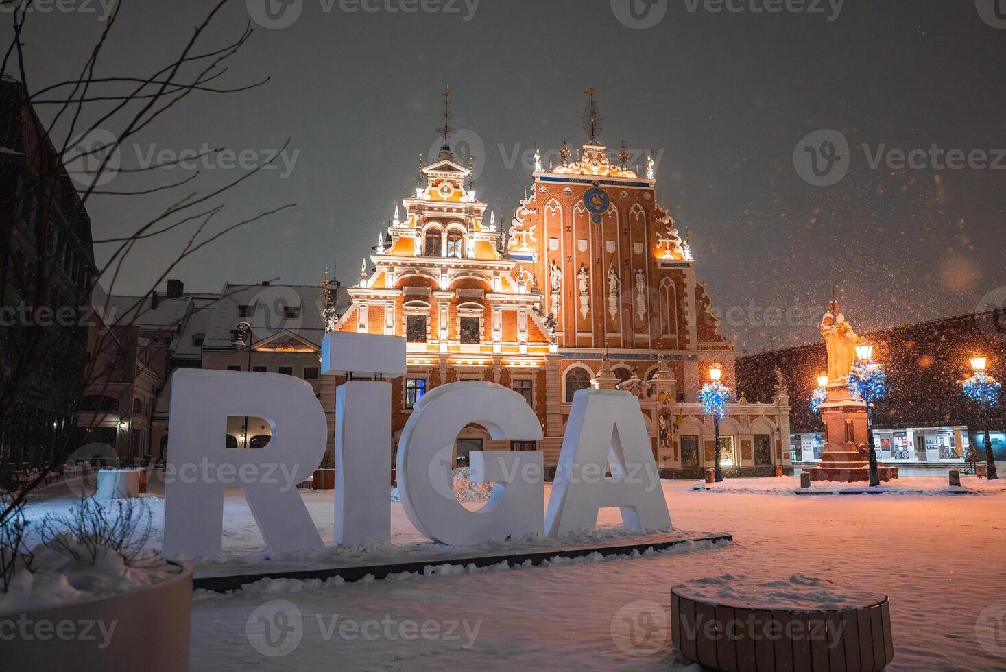 The House of the Blackheads in Riga, Latvia, glows under winter lights with snow covered ground, blue lit trees, and a statue in the square. photo