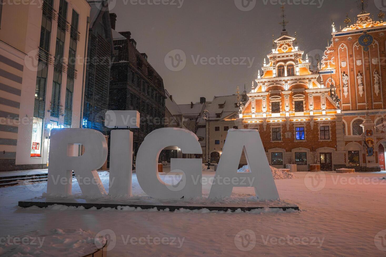 A snowy evening in Riga, Latvia, featuring the RIGA sign, the illuminated House of the Blackheads, and surrounding buildings in a serene winter setting. photo