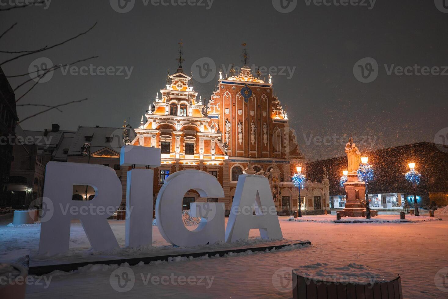 Winter evening in Riga, Latvia, with the illuminated House of the Blackheads, snow covered ground, streetlights, and a statue in a festive square. photo