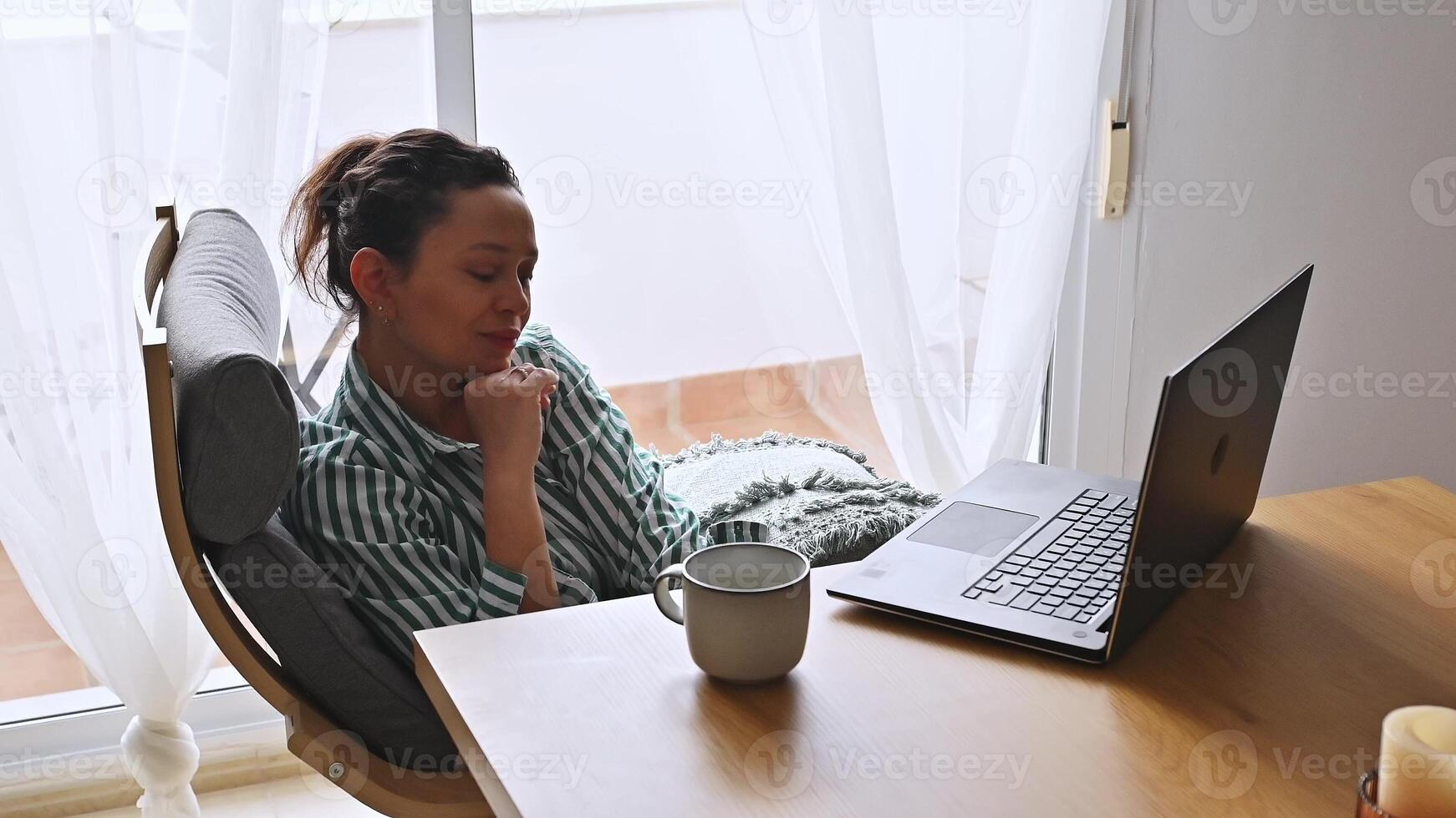 A young woman enjoying remote work from her comfortable home office setting. She sits with a laptop and coffee, exuding serenity and productivity in a bright, inviting room. photo