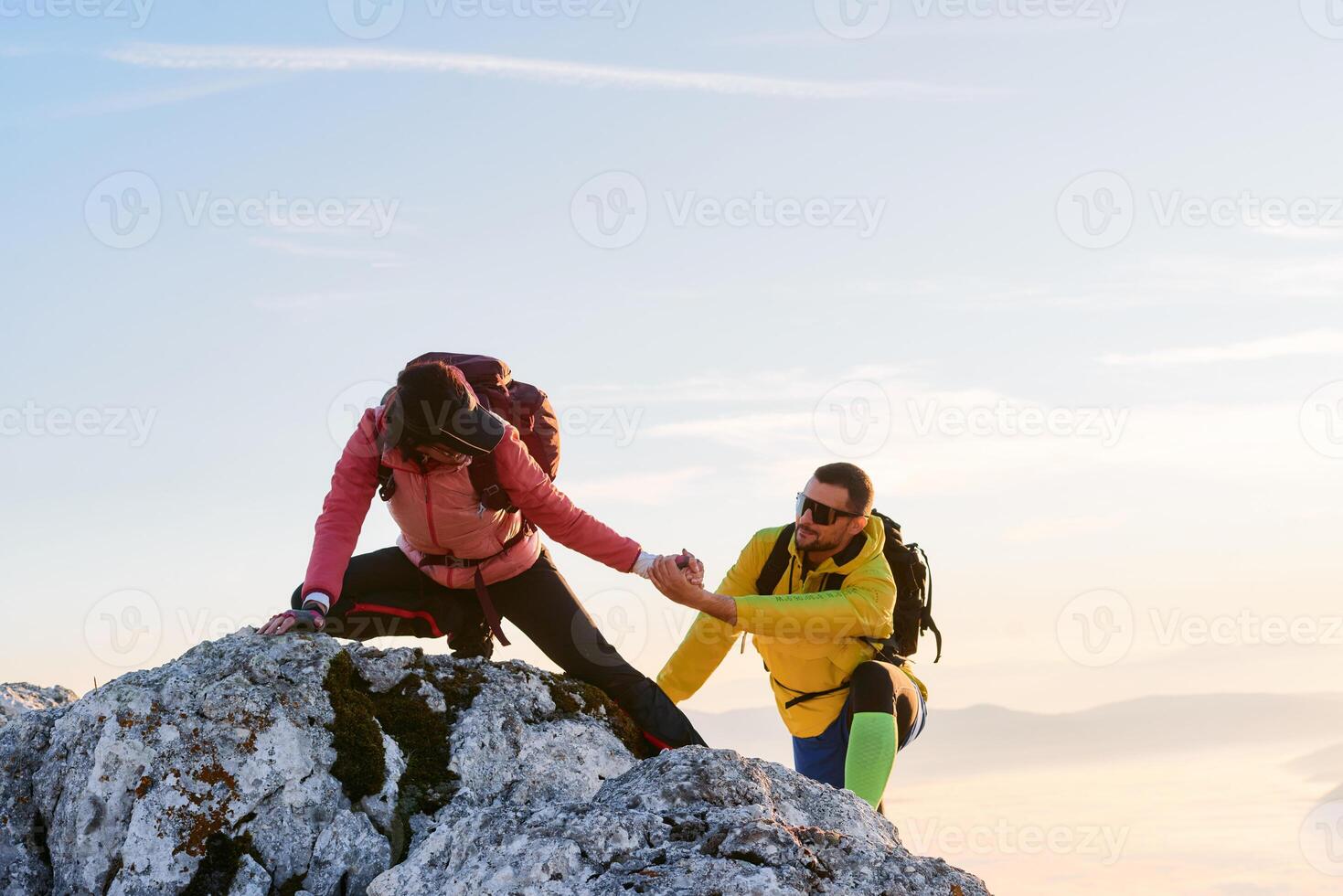 Two hikers, couple in outdoor gear climb rocky terrain with the sun rising behind them.Climbers reaching the mountain peak at sunrise. photo