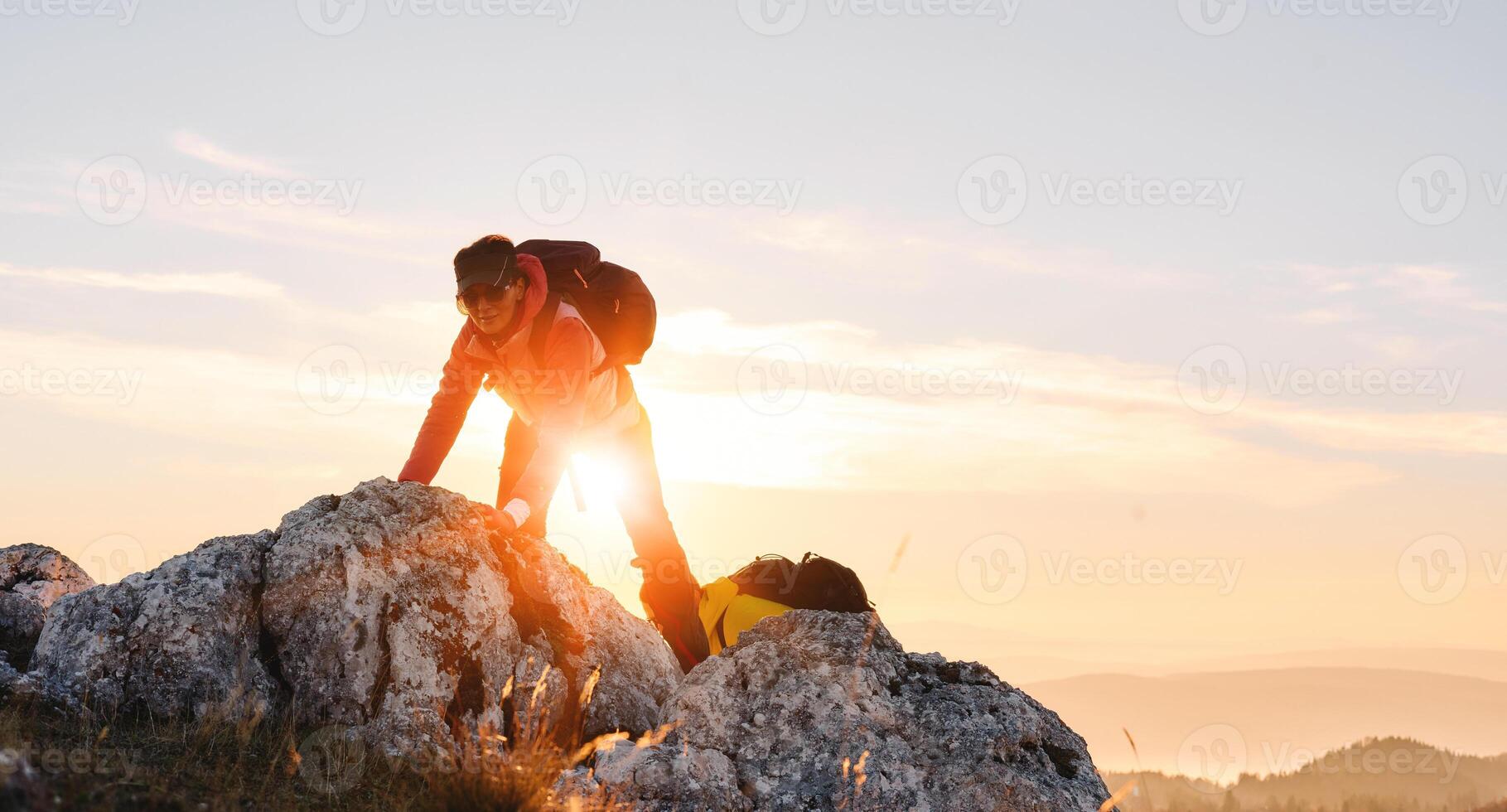 Two hikers, couple in outdoor gear climb rocky terrain with the sun rising behind them.Climbers reaching the mountain peak at sunrise. photo
