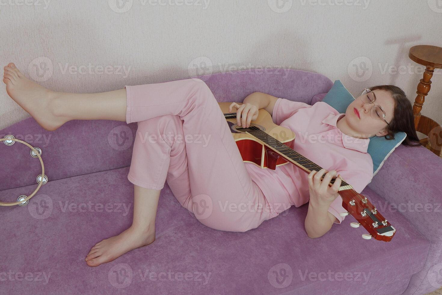 Woman guitarist with eyeglasses and her eyes closed laying on her back on sofa and playing guitar. Musician of Caucasian ethnicity dressed casually in jeans, polo shirt. High angle view photo