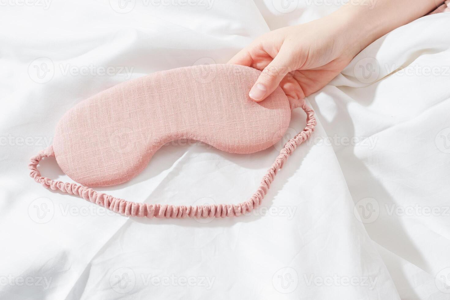 Close up Pink eye mask for sleep in woman hand on white bedclothes, minimal lifestyle aesthetic flat lay. Top view sleeping mask for best sleepers, for travel, comfort relax. Rest well concept. photo