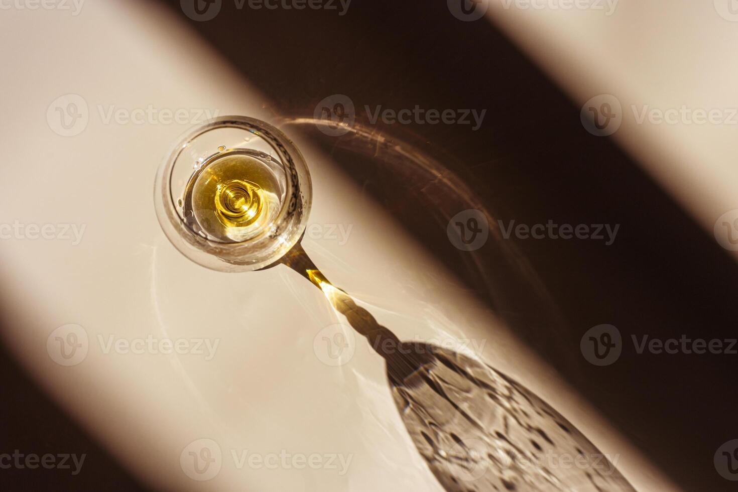 One glass of cognac sparkling at sunlight, beige background with dark shadows and reflection, sun flare light effect on table. Strong alcohol drink with star filter, top view, desaturated colors photo