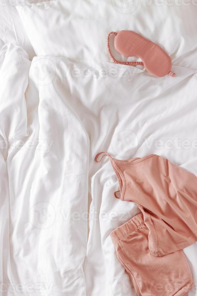 Top view pink pajama and eye sleep mask on white crumpled bedclothes, cushion and blanket. Cozy pyjamas for comfort rest at night. Flat lay from singlet, shorts, sleeping mask pastel pink color photo