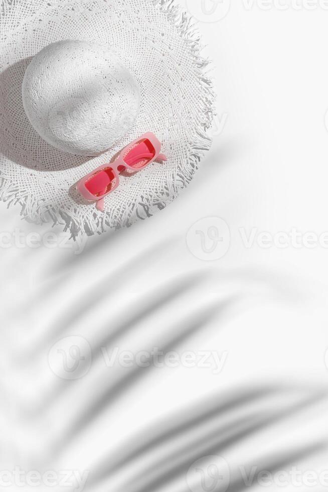 White sun hat and pink colored sunglasses on white background at sunlight with palm leaf shadows. Summer vacation, summertime relaxation concept, minimal trend flat lay, copy space. photo