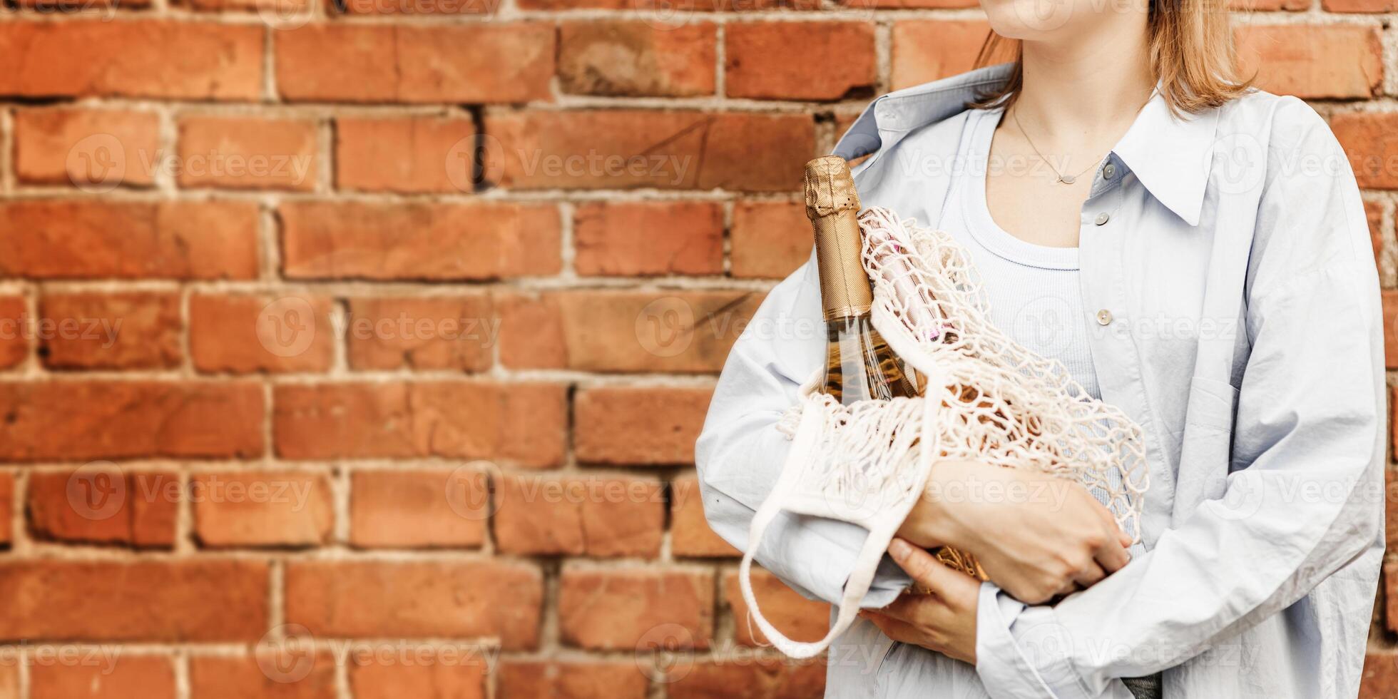 Girl holding champagne sparkling white and rose wine in hands, standing near brick wall, no face fashion style, holiday lifestyle photo. Female wearing casual clothes on street with alcohol photo