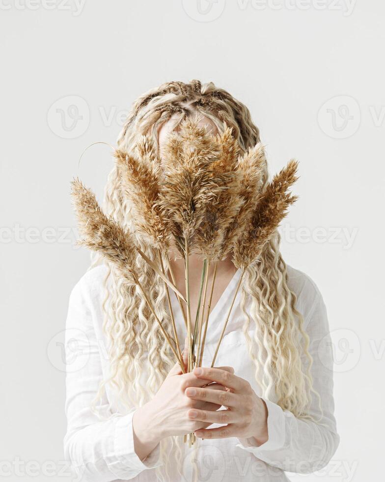Woman holding pampas grass in front of face, light background, no face trend concept. Female with long curly hair dreadlocks hiding behind bouquet dry flower. Creative autumn visual. Fall concept photo